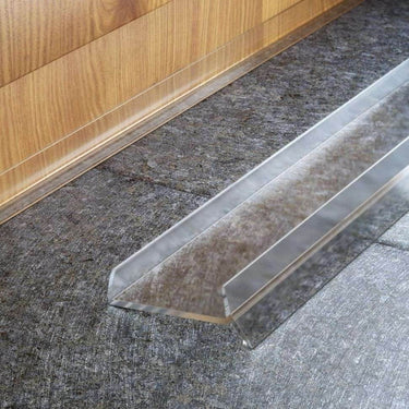 This image shows a transparent PVC plinth sealing strip fitted to a kitchen base unit kickboard. The strip protects the bottom of the wooden plinth from water, dirt, and damage, creating a neat, moisture-resistant barrier where the floor meets the cabinet.
